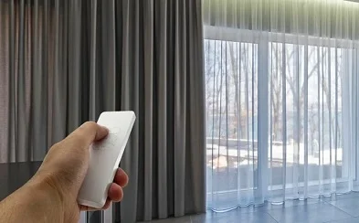 Hand using remote control to operate motorized gray curtains and sheers
